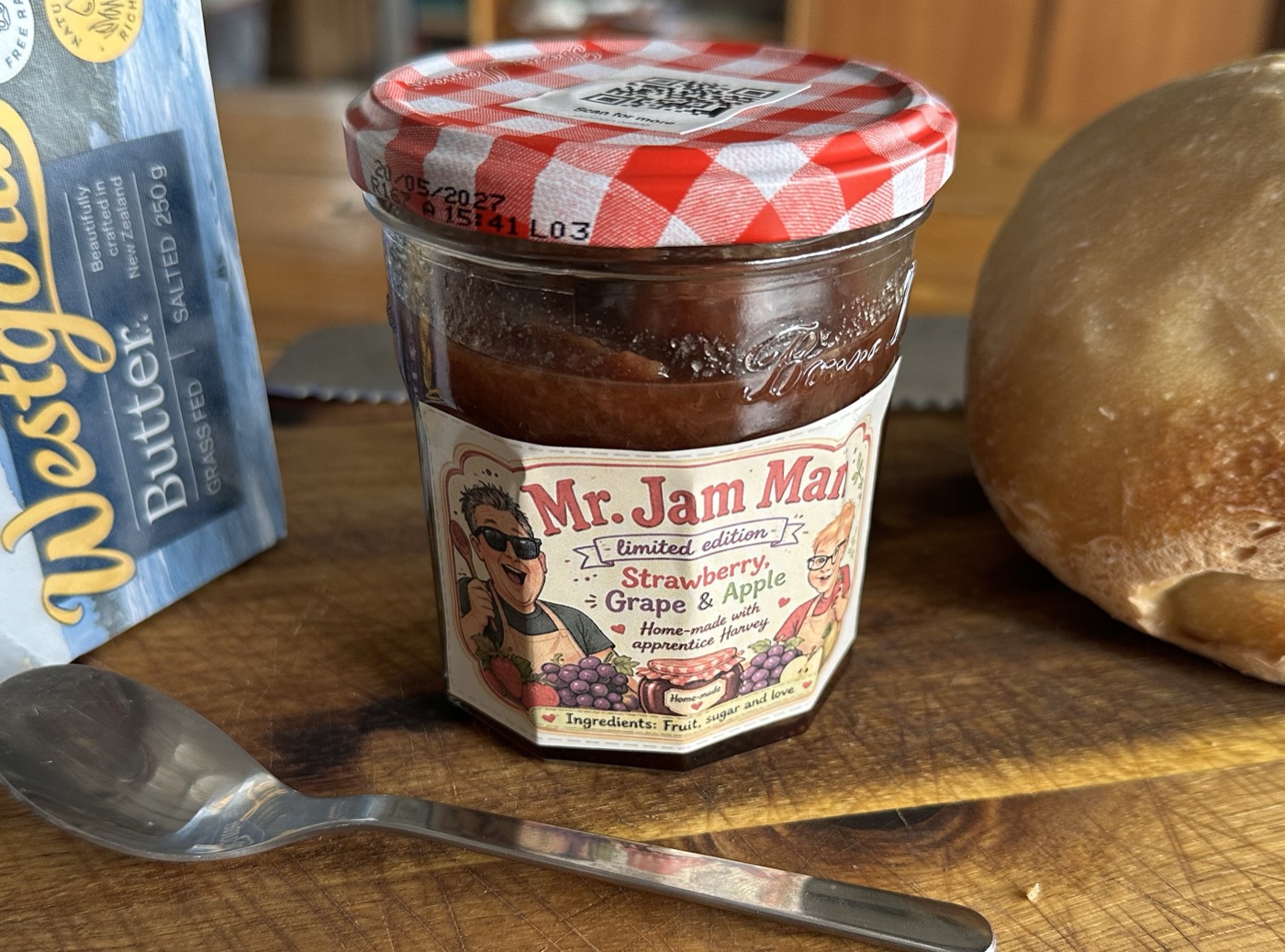 A jar of Mr. Jam Man strawberry, grape and apple jam on a wooden board, with a teaspoon, a loaf of bread and a block of butter